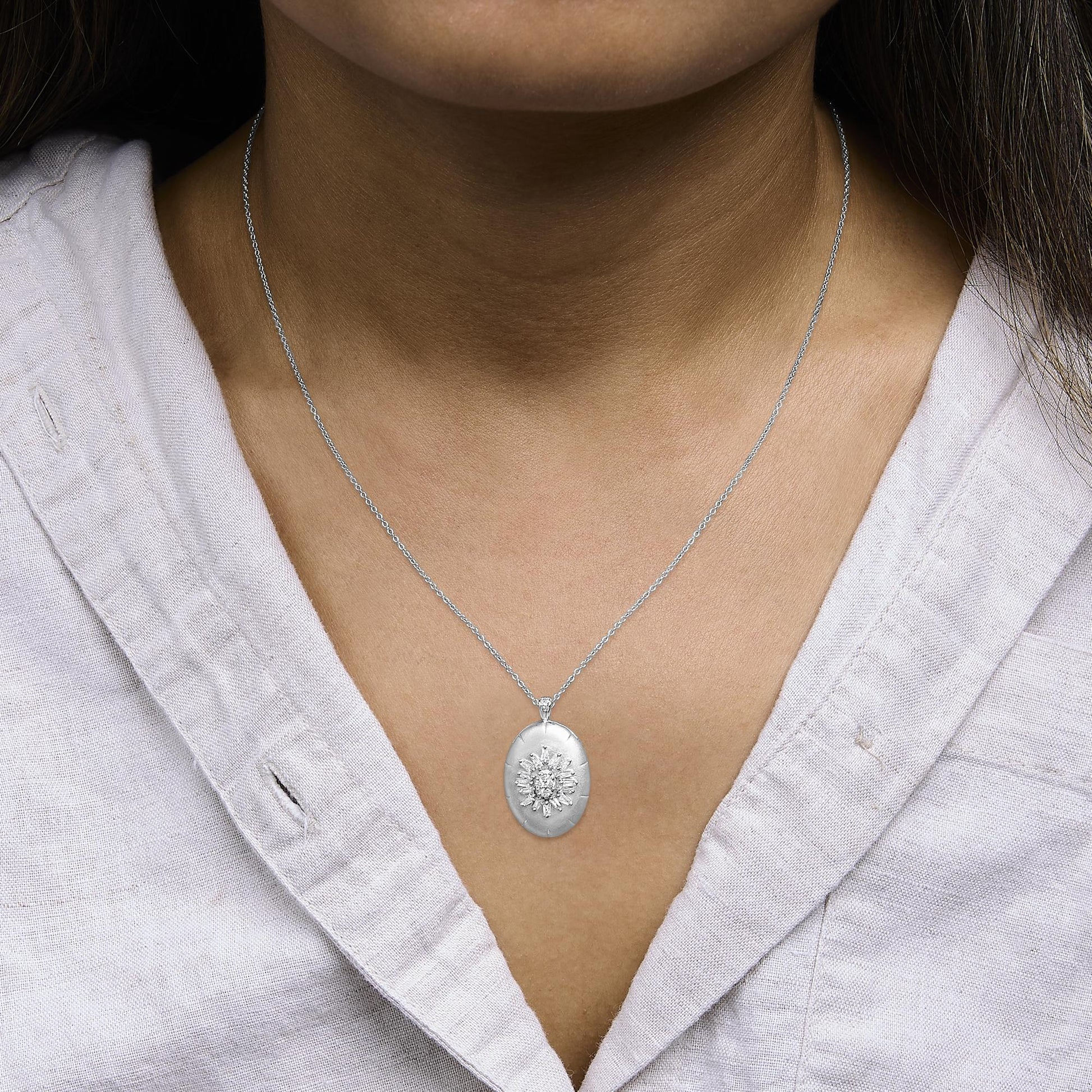White metal necklace shown worn on a model, featuring an oval pendant with round and baguette-shaped white gemstones in prong settings, photographed from a front close-up angle.