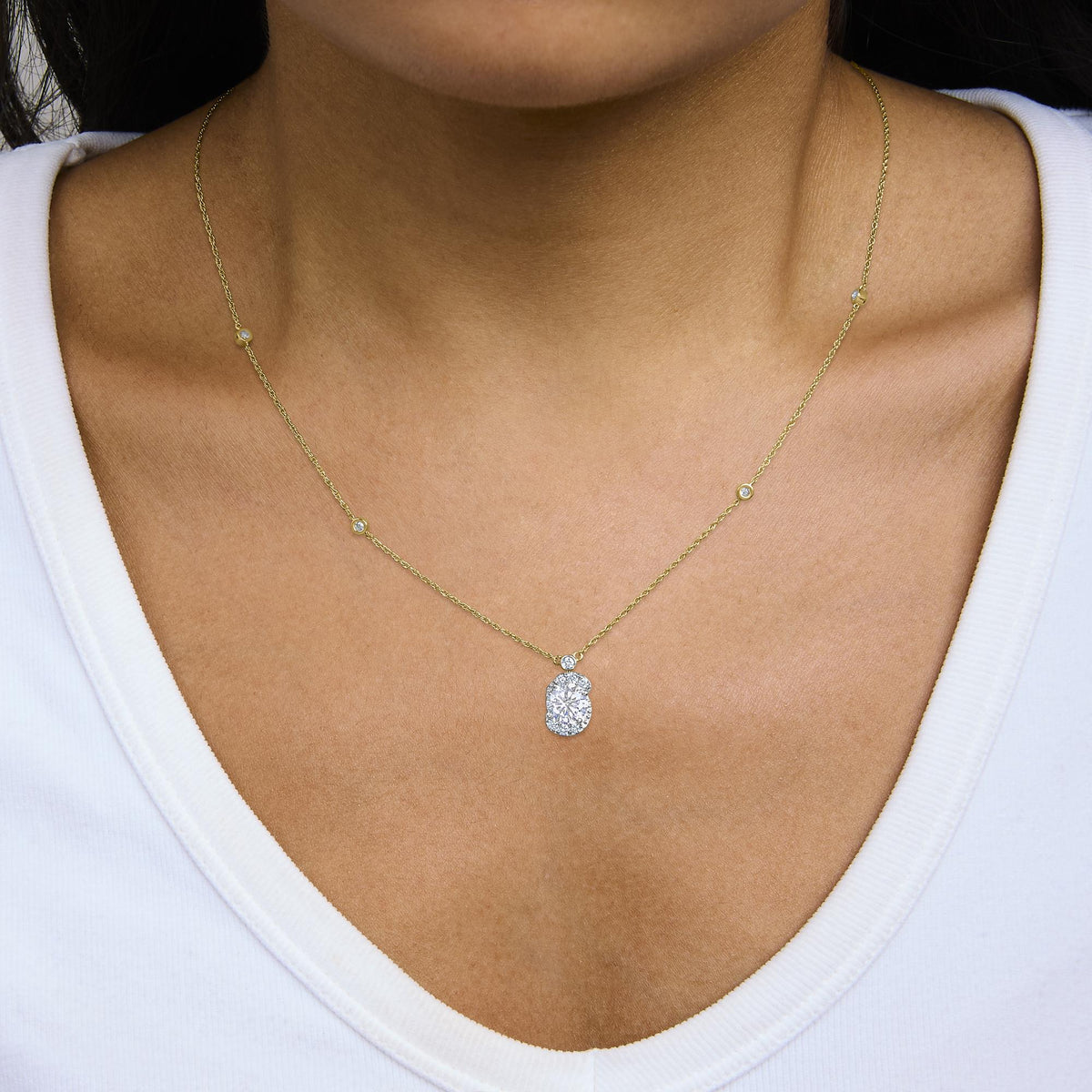 Close-up front view of a model wearing a yellow metal necklace with a central pendant featuring a cluster of white gemstones surrounded by smaller white gemstones. The chain includes bezel-set white gemstones spaced along it. The model is wearing a white top.
