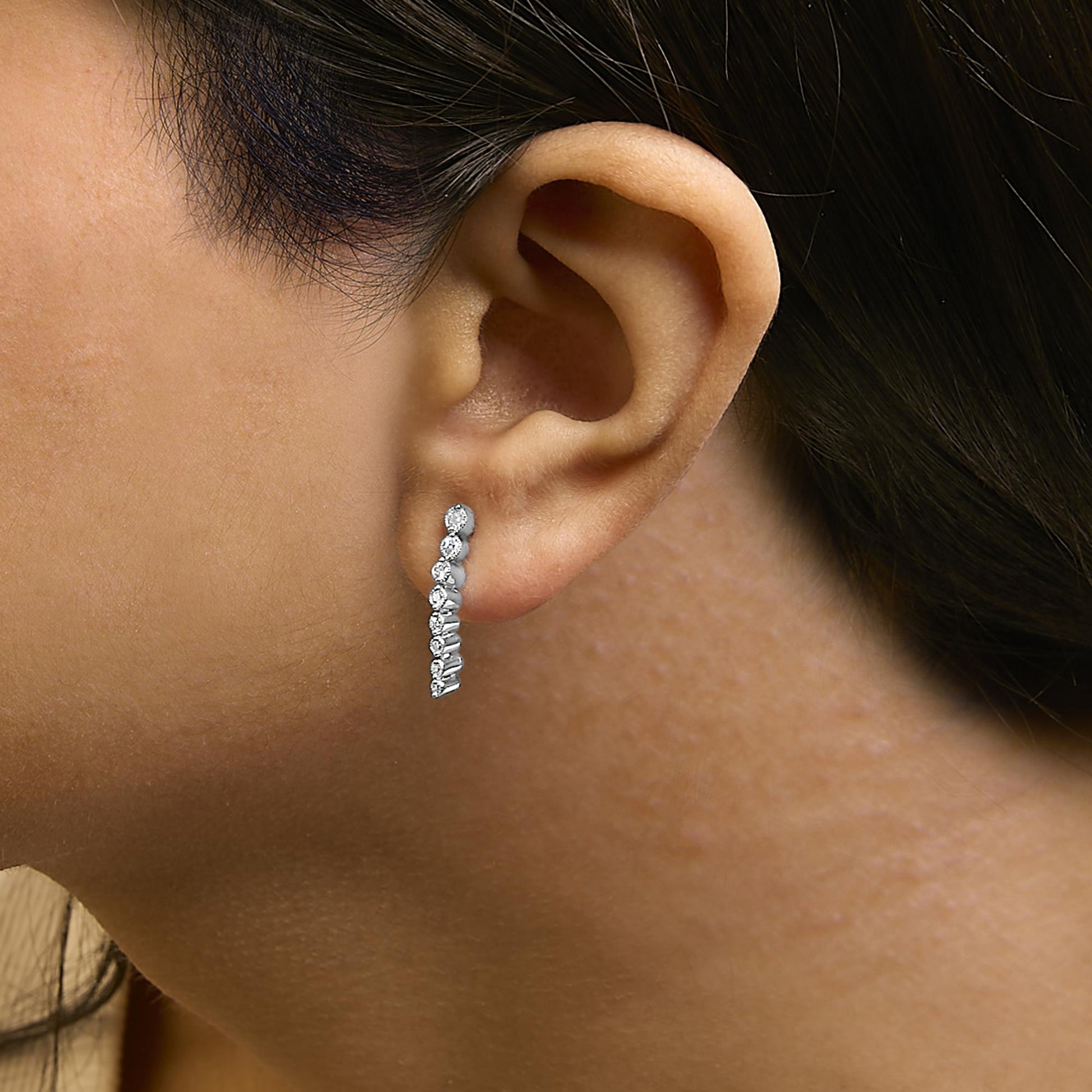 Close-up side view of a model wearing a white metal earring with a vertical row of round white gemstones in prong settings.