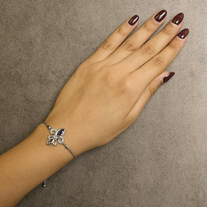 White metal bracelet with a fleur-de-lis design featuring small round white gemstones and two marquise-shaped bezel-set purple and orange gemstones, shown on a model's wrist from a top-down angle.