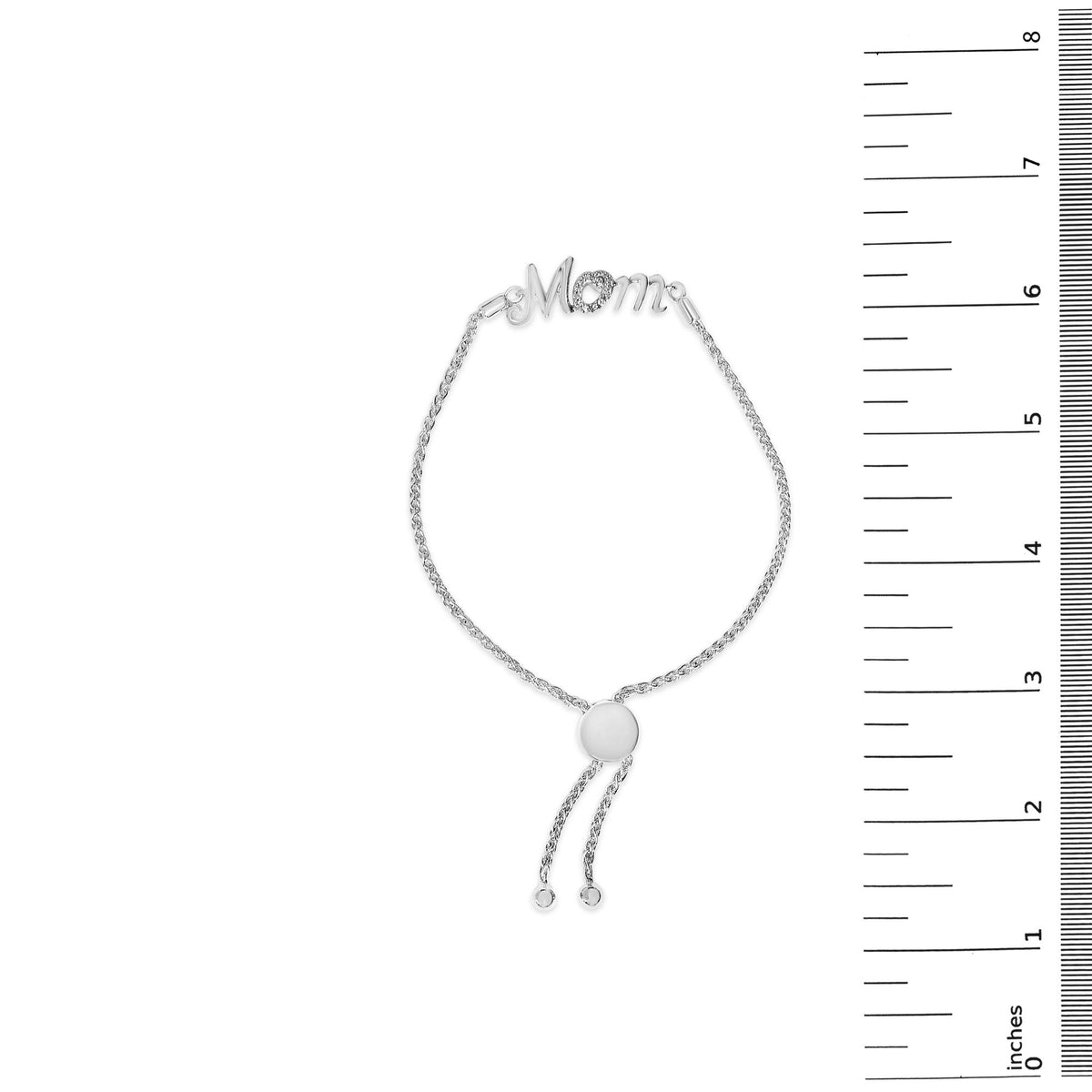 White bracelet with 'Mom' in script, the 'o' is heart shaped and set with white gemstones in pave setting, shown on a ruler scale, top view.