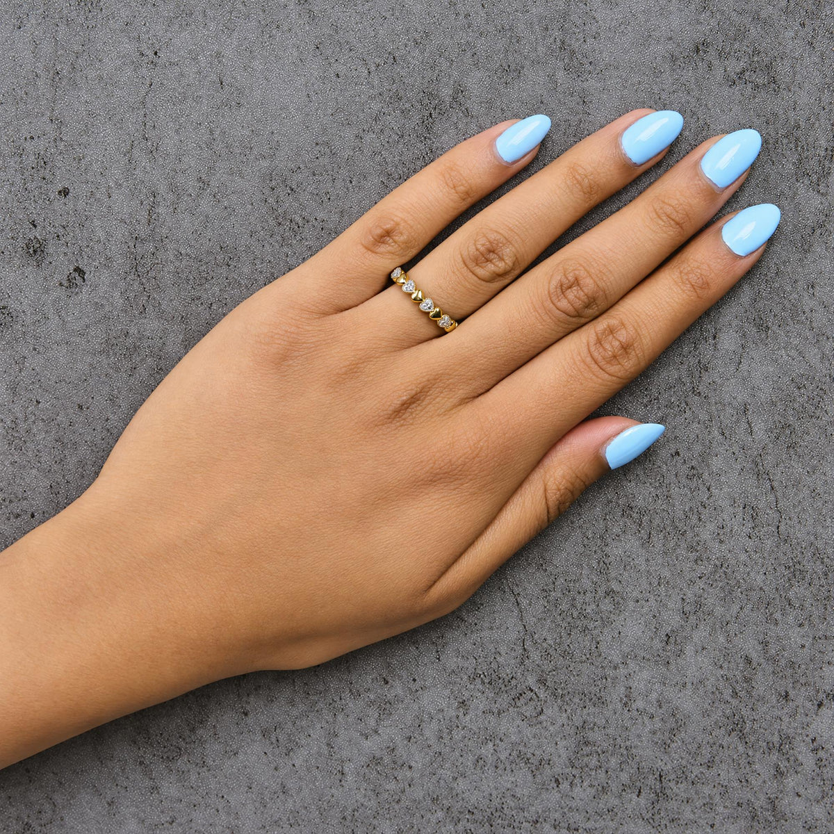 Yellow metal ring with alternating heart shapes and white gemstones in pave setting, shown worn on a model's finger, top-down angle.