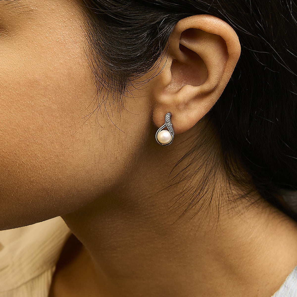 Close-up side view of a model wearing white earrings with a round pearl and curved design featuring round white gemstones in pave setting.