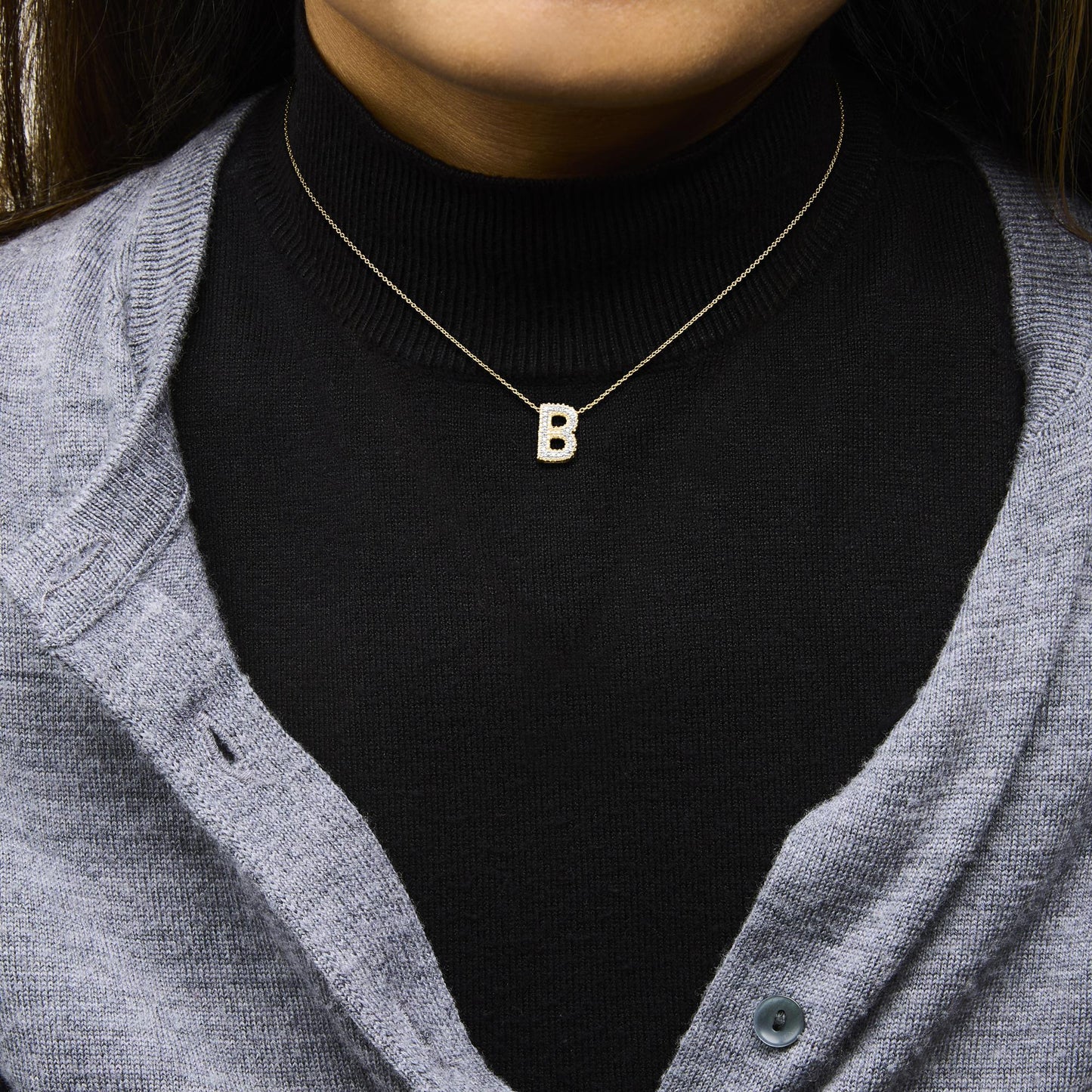 Close-up front view of a model wearing a yellow metal necklace with a pendant shaped like the letter 'B' encrusted with prong-set round white gemstones.