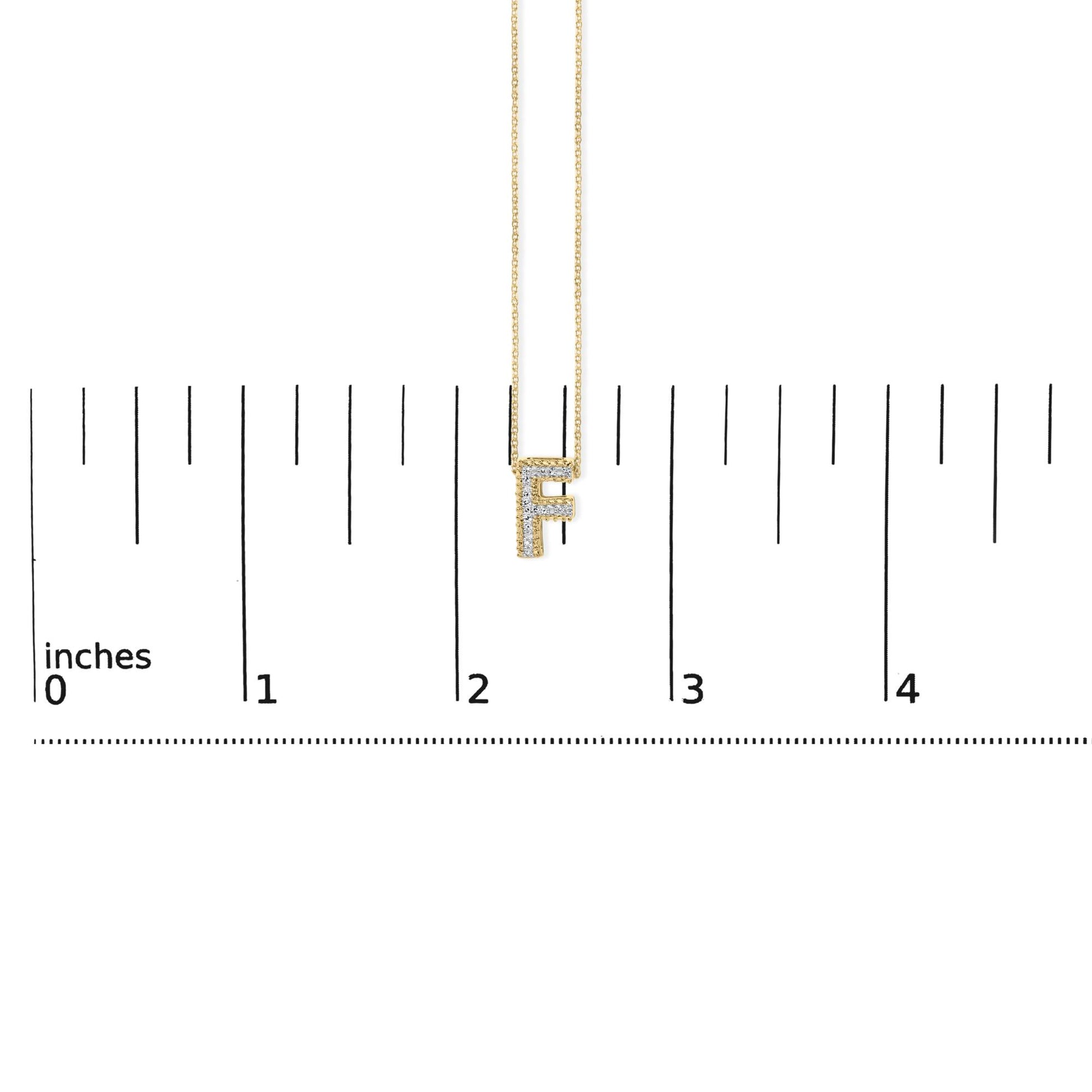 Close-up front view of a yellow metal necklace pendant in the shape of the letter 'F' set with multiple white gemstones, shown hanging on a chain and placed on a ruler scale background.