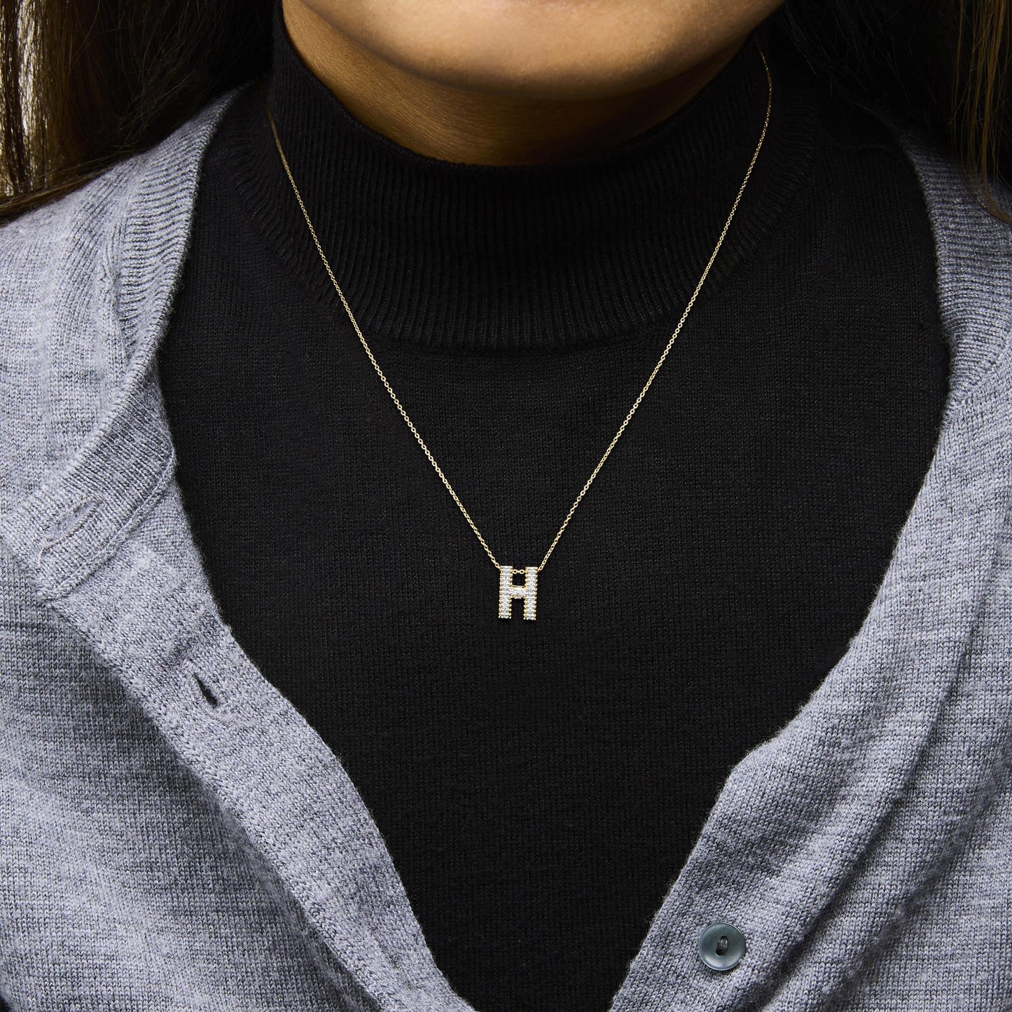 Close-up front view of a necklace with a yellow chain and a pendant in the shape of the letter 'H' adorned with white gemstones, shown worn on a model dressed in a black turtleneck and grey cardigan.