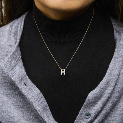 Close-up front view of a necklace with a yellow chain and a pendant in the shape of the letter 'H' adorned with white gemstones, shown worn on a model dressed in a black turtleneck and grey cardigan.