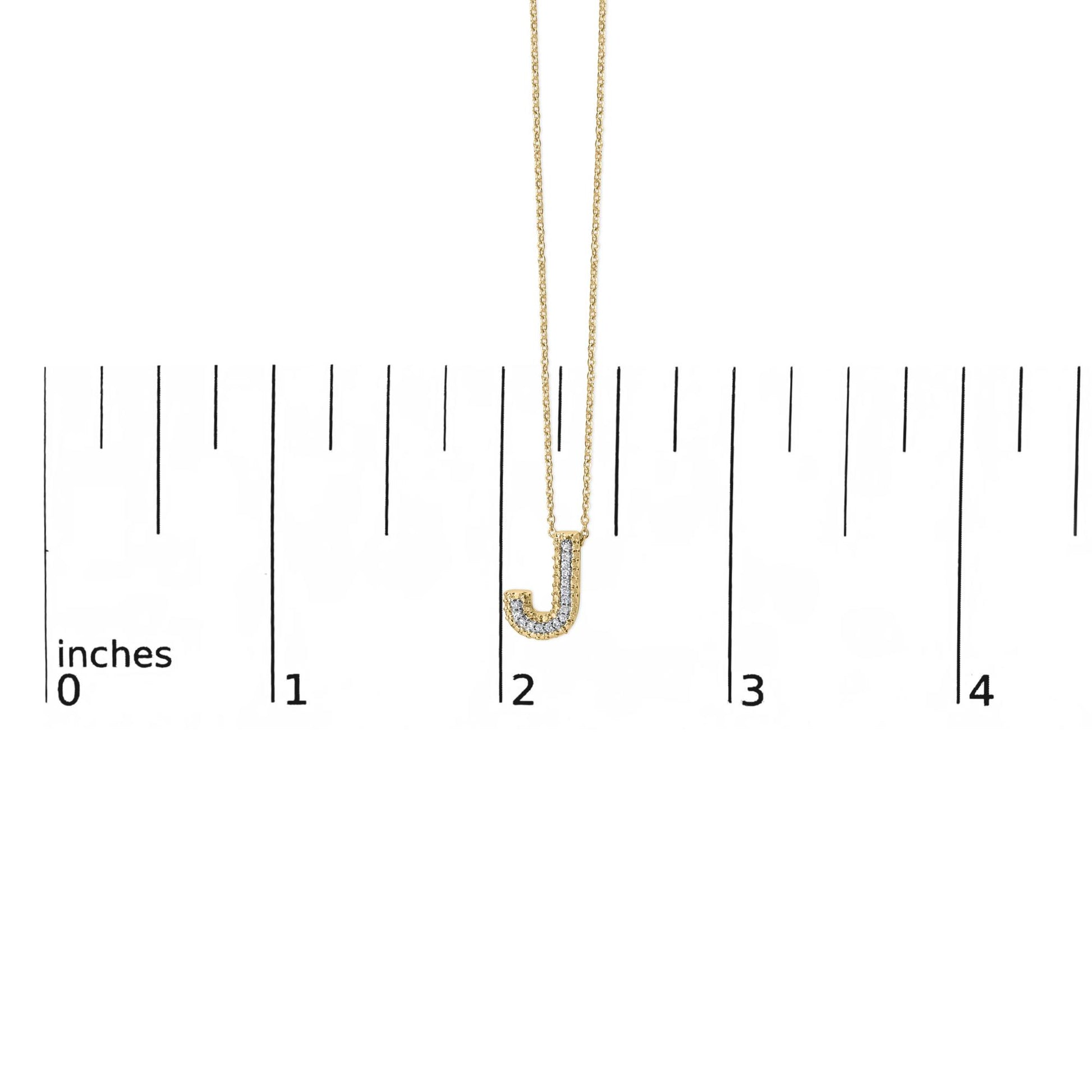 Close-up front view of a yellow metal necklace with a pendant in the shape of the letter J, featuring white round gemstones in a prong setting, shown on a ruler scale.