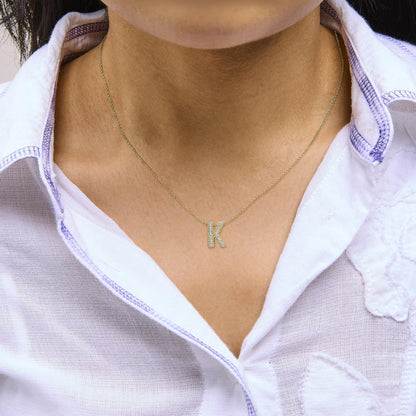 Close-up front view of a model wearing a yellow metal necklace with a pendant shaped as the letter 'K,' adorned with multiple white gemstones in prong settings.