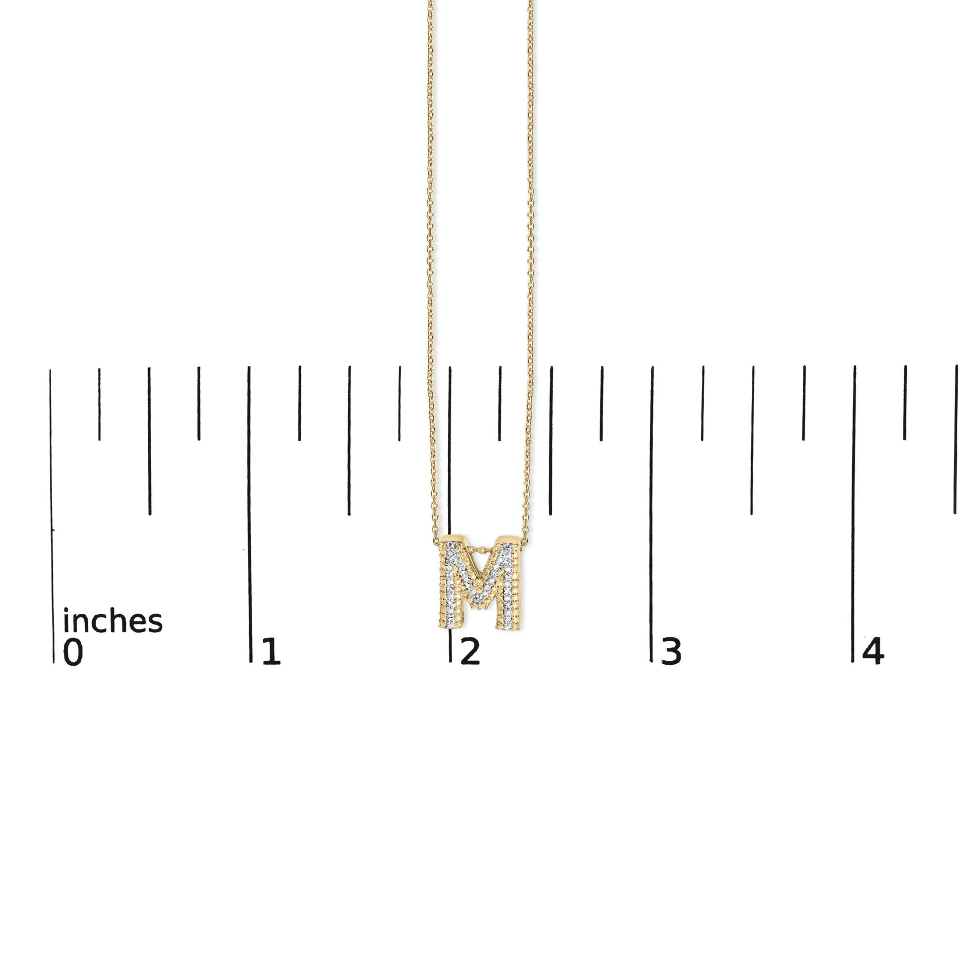 Close-up front view of a yellow metal necklace with an 'M' pendant adorned with white round gemstones in prong settings, shown on a scale.
