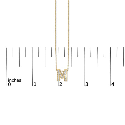 Close-up front view of a yellow metal necklace with an 'M' pendant adorned with white round gemstones in prong settings, shown on a scale.