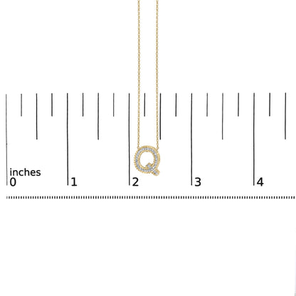 Close-up angled front view of a yellow metal necklace pendant in the shape of the letter Q, outlined with white gemstones in prong settings, shown on a ruler scale.