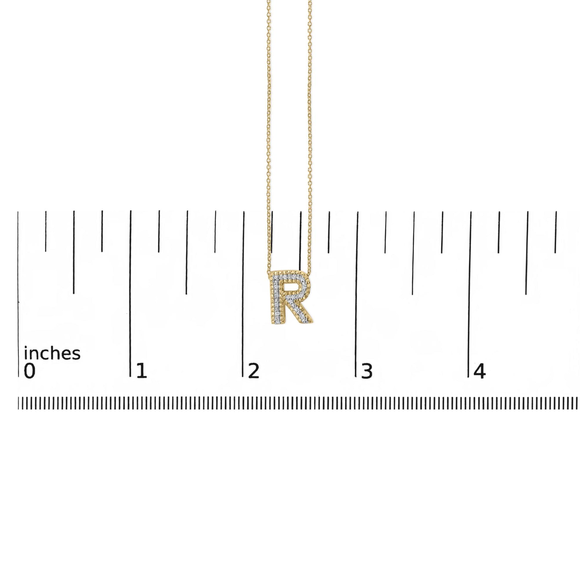 Image of a yellow metal necklace pendant shaped as the letter 'R' adorned with white gemstones in a prong setting, shown hanging on a chain against a ruler scale, viewed from the front.
