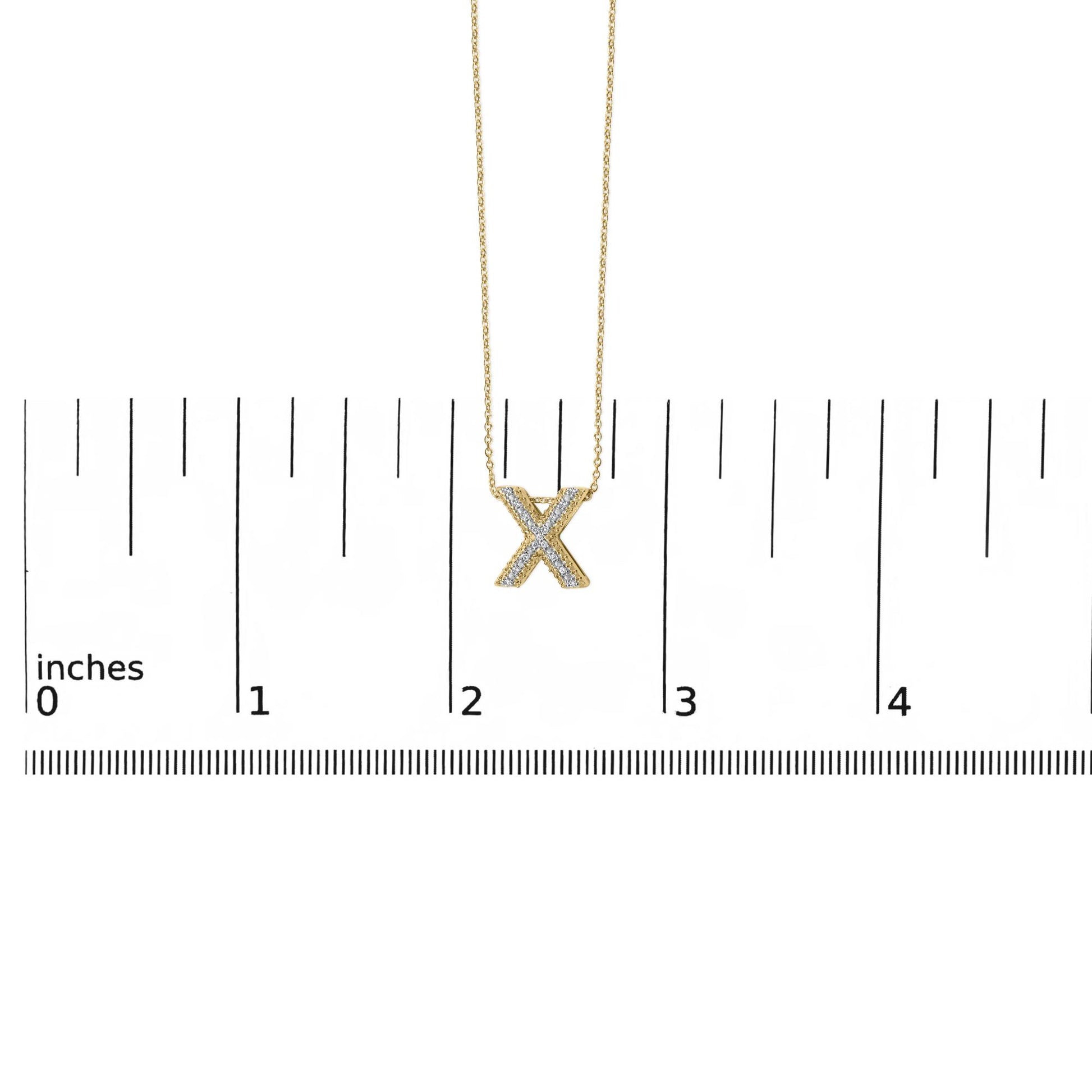 Yellow metal necklace with an X-shaped pendant featuring white round gemstones in a prong setting, shown hanging against a ruler scale, front view.