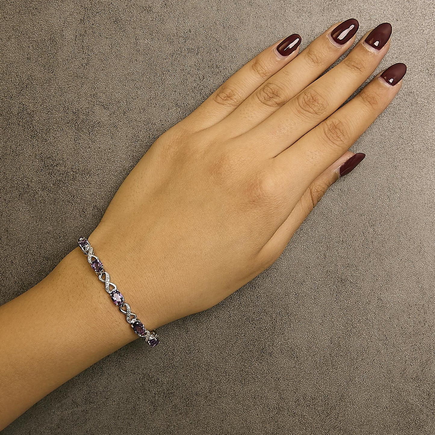White metal bracelet with alternating oval purple gemstones and infinity-shaped links with white gemstones, shown on a model's wrist at top angle.