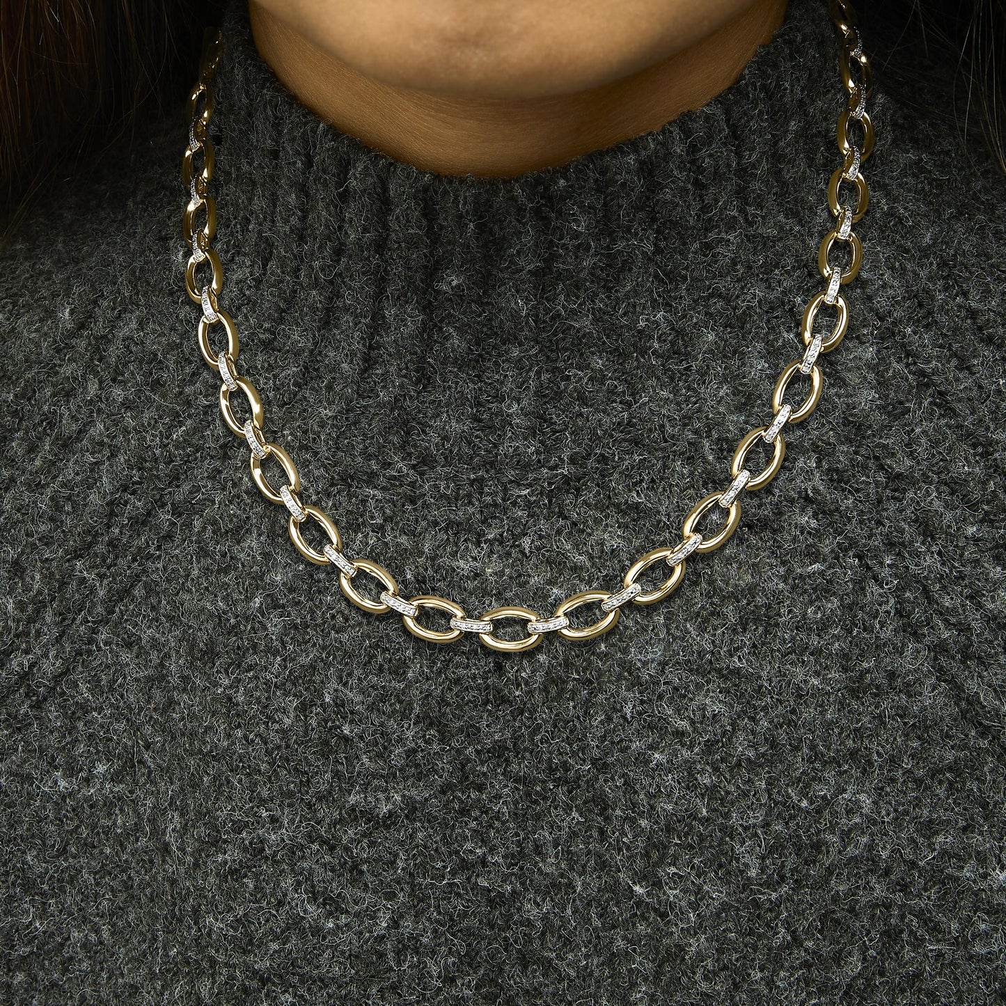 Close-up front view of a yellow metal necklace with large oval links, some links set with white gemstones in pave setting, worn on a model with a dark textured sweater.