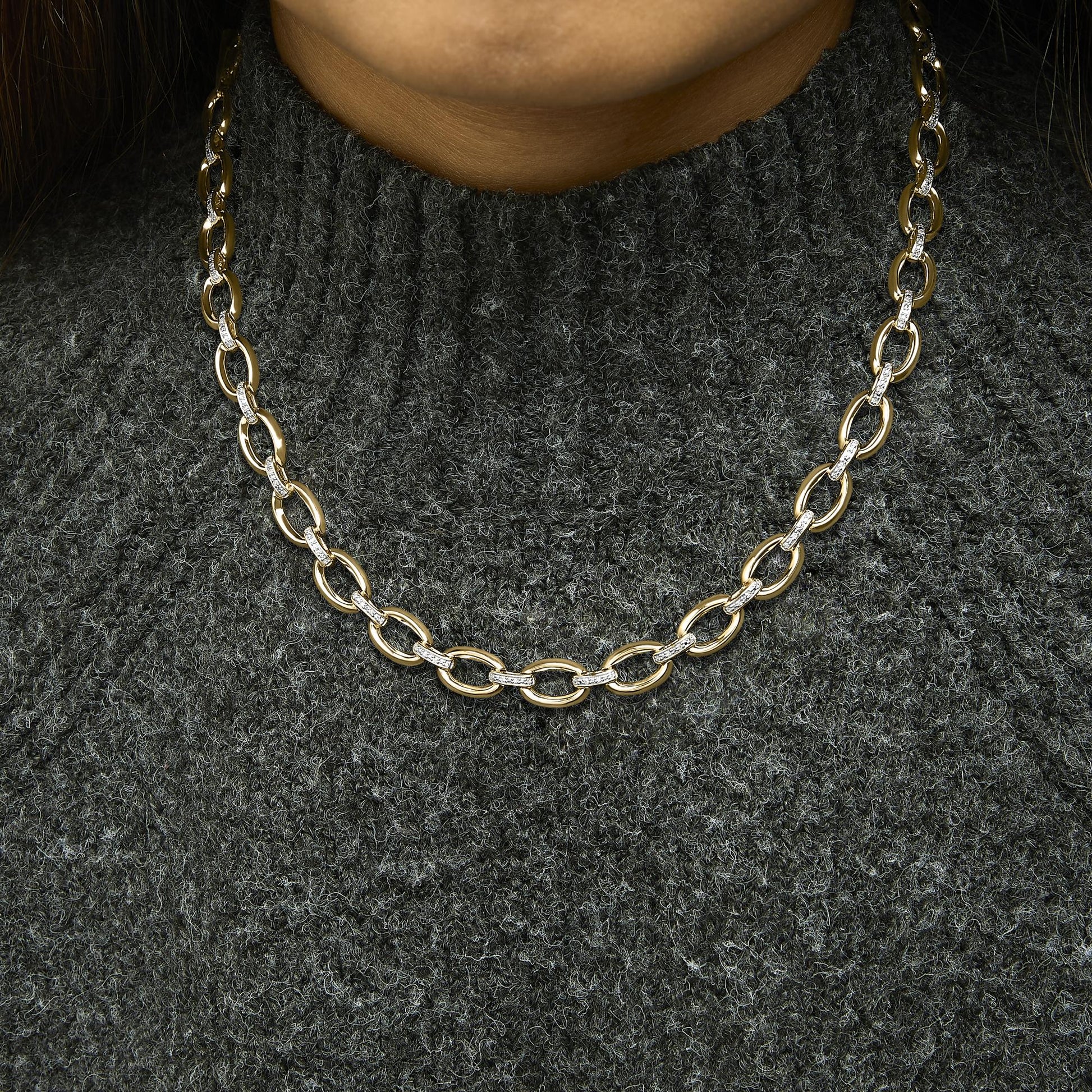 Close-up front view of a yellow metal necklace with large oval links, some links set with white gemstones in pave setting, worn on a model with a dark textured sweater.