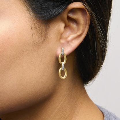 Close-up side view of a model wearing yellow metal drop earrings with interlocking oval shapes and vertical bars adorned with white pave-set round gemstones.