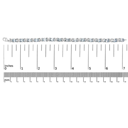 Top-down view of a white metal bracelet with alternating oval blue gemstones and round white gemstones in prong settings, placed on a ruler for size reference.