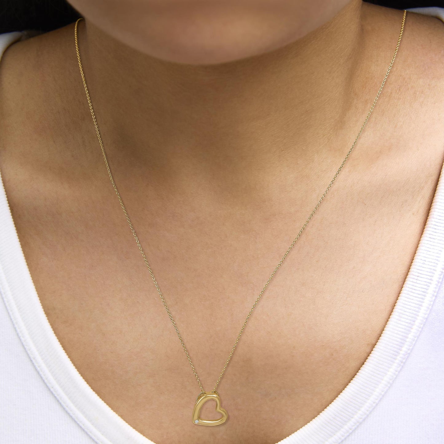 Yellow metal heart-shaped pendant necklace with a small white gemstone in the lower left corner of the pendant, shown worn on a model, close-up front angle.