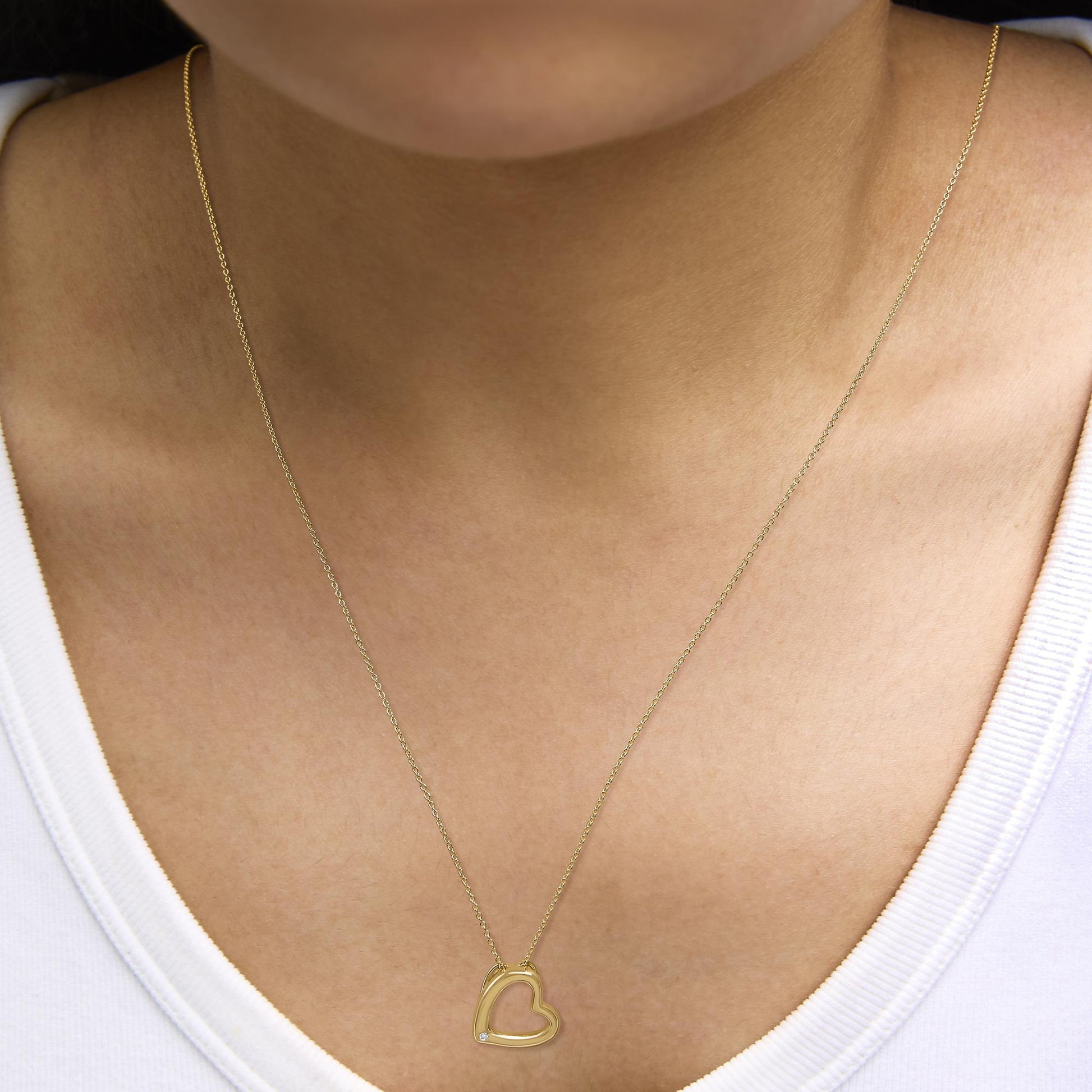 Yellow metal heart-shaped pendant necklace with a small white gemstone in the lower left corner of the pendant, shown worn on a model, close-up front angle.