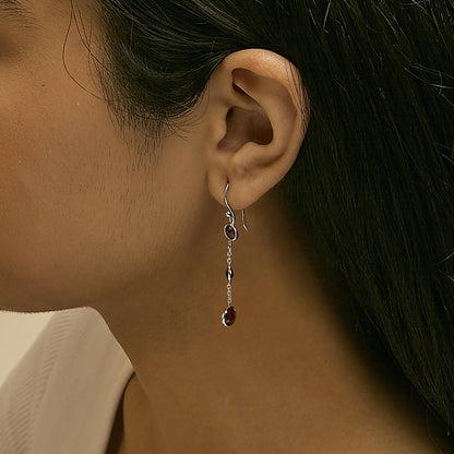 Close-up side view of a model wearing a pair of white metal dangle earrings featuring multiple round red gemstones in bezel settings connected by chains.