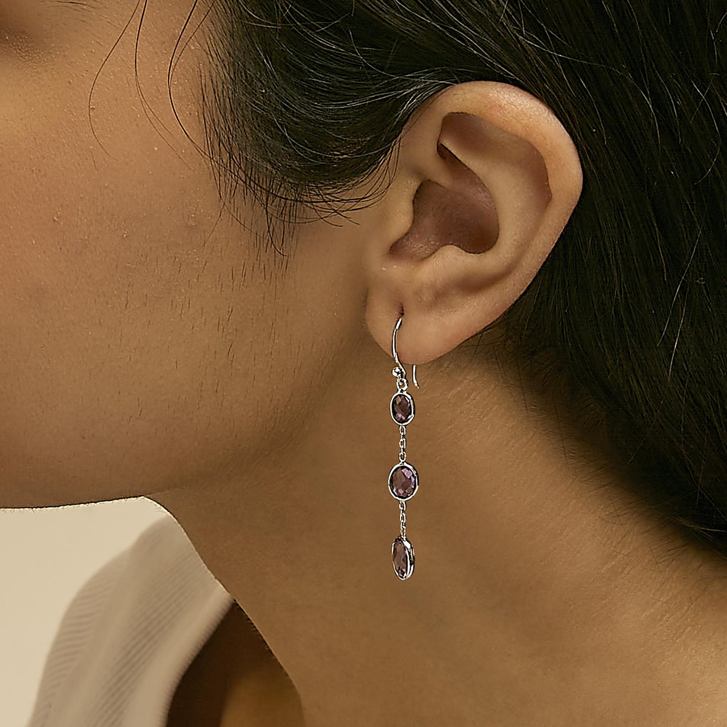 Side view of a white metal dangle earring worn on a model, featuring three linked oval-shaped purple gemstones in bezel settings.