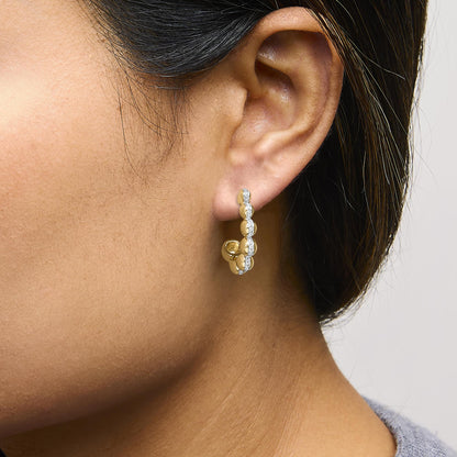 Close-up side view of a model wearing yellow metal hoop earrings with a vertical row of white gemstones in a pave setting.