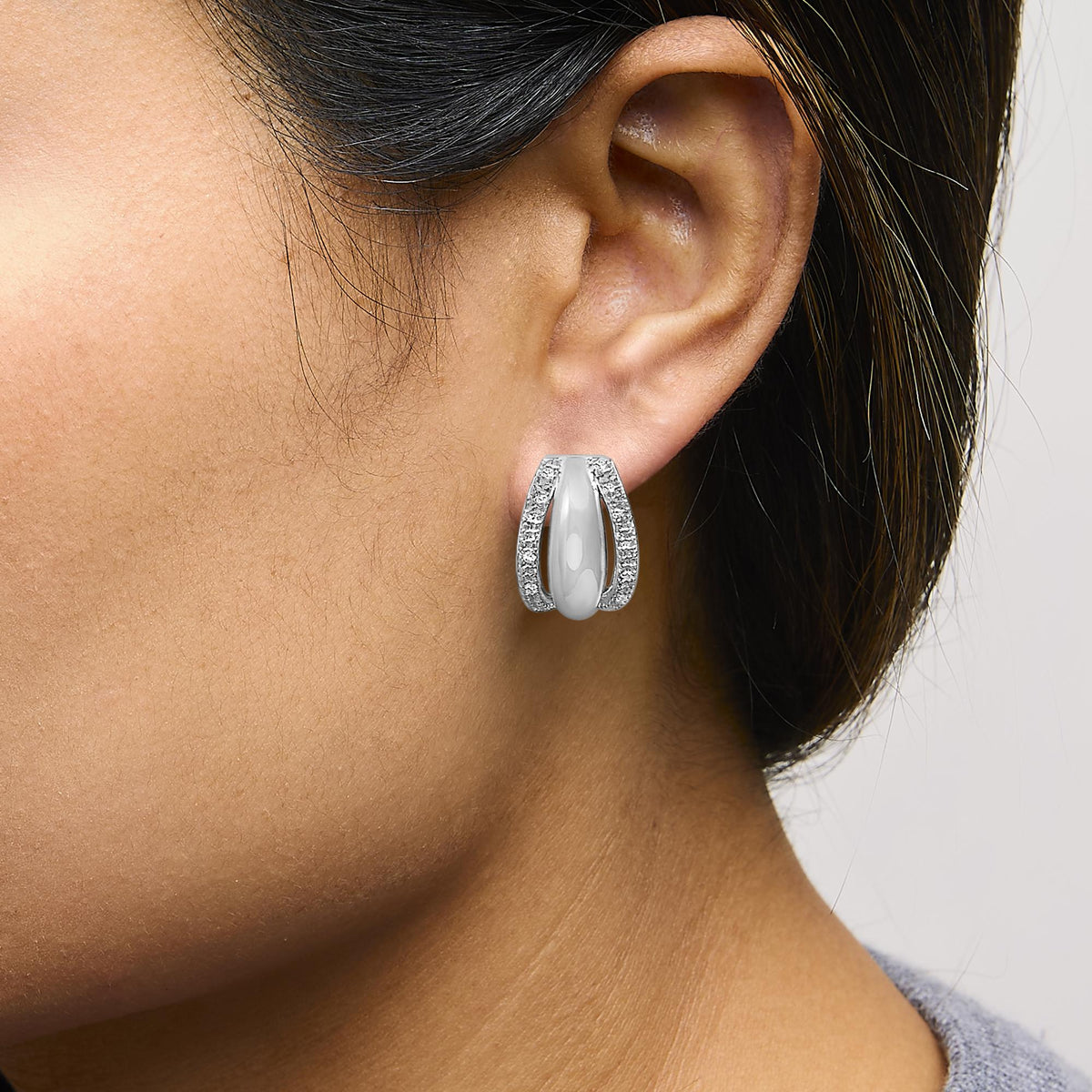 Close-up front angle of a model wearing white metal earrings with pave-set round white gemstones outlining an elongated oval shape.