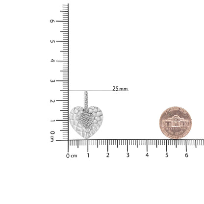 Close-up front view of a white metal heart-shaped necklace pendant with a textured outer heart and a smaller heart in the center set with white round pave gemstones, placed on a ruler and next to a coin for size reference.