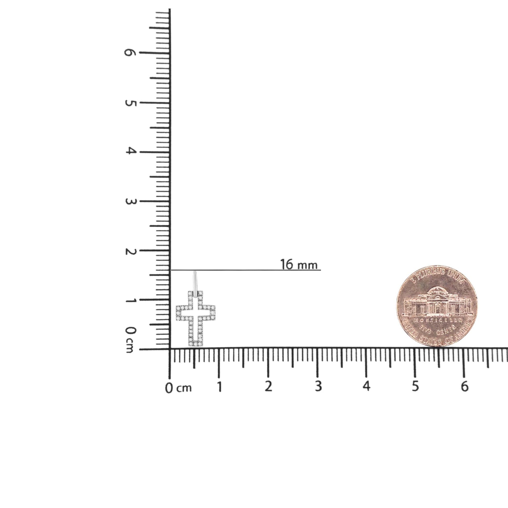 Close-up front view of a white metal cross-shaped necklace pendant with round white gemstones in a pave setting, placed on a ruler and next to a coin for size reference.