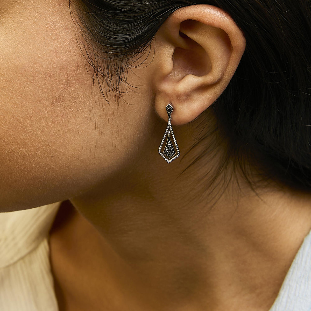 Close-up side view of a model wearing white metal drop earrings with a geometric design featuring black gemstones in a pave setting.