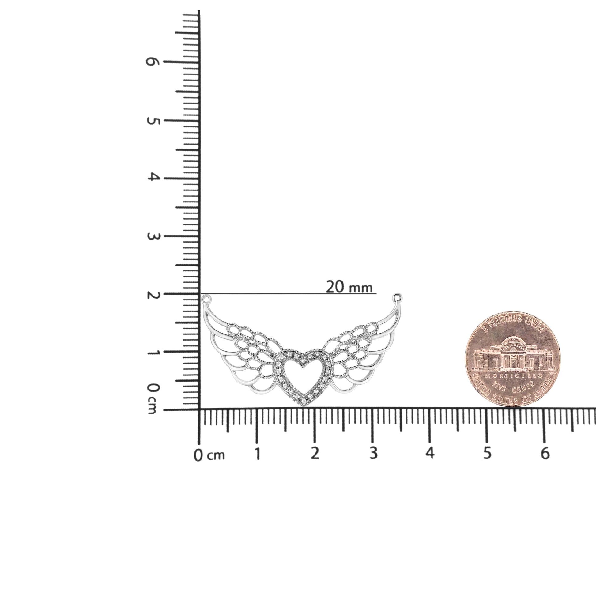 Top-down view of a white metal necklace pendant with a heart shape in the center surrounded by pave-set round white gemstones and wing-like designs on each side, placed on a ruler and a coin for size reference.