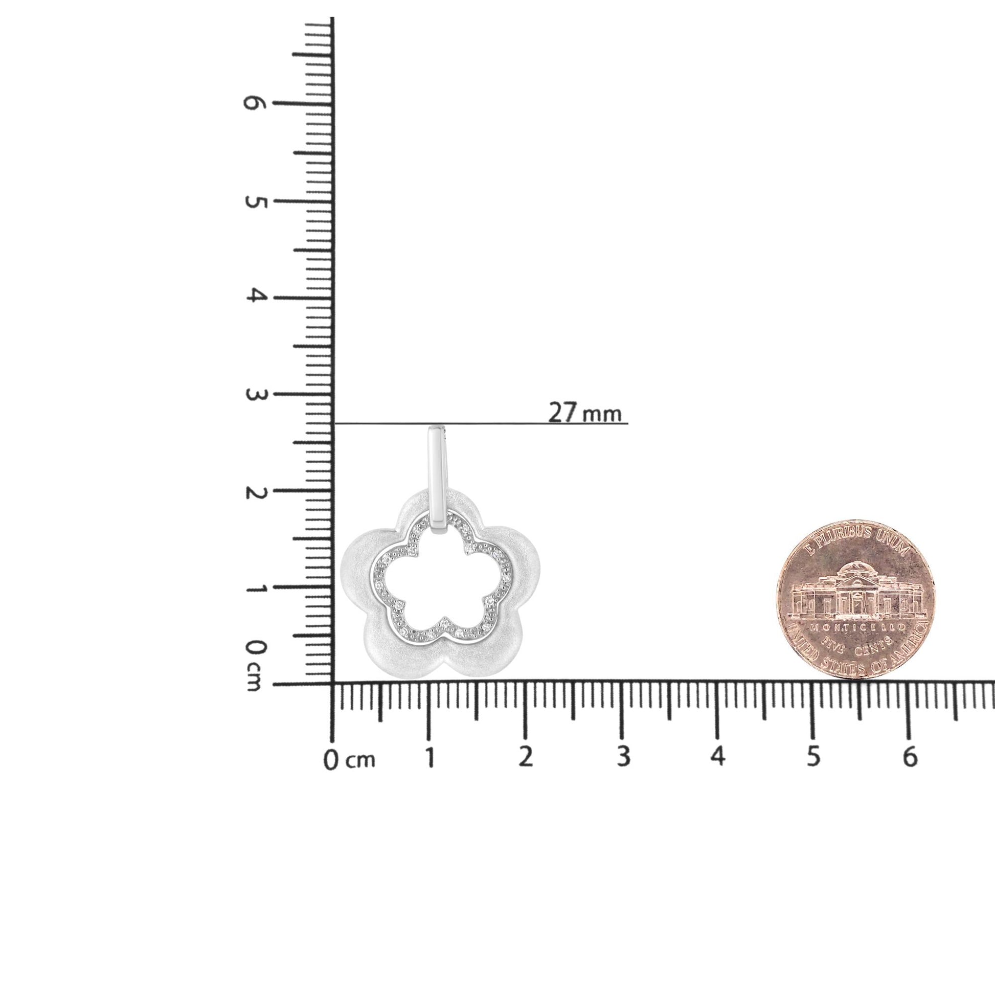 White metal flower-shaped necklace pendant with pave set round white gemstones, shown from front on a ruler and coin for size reference.