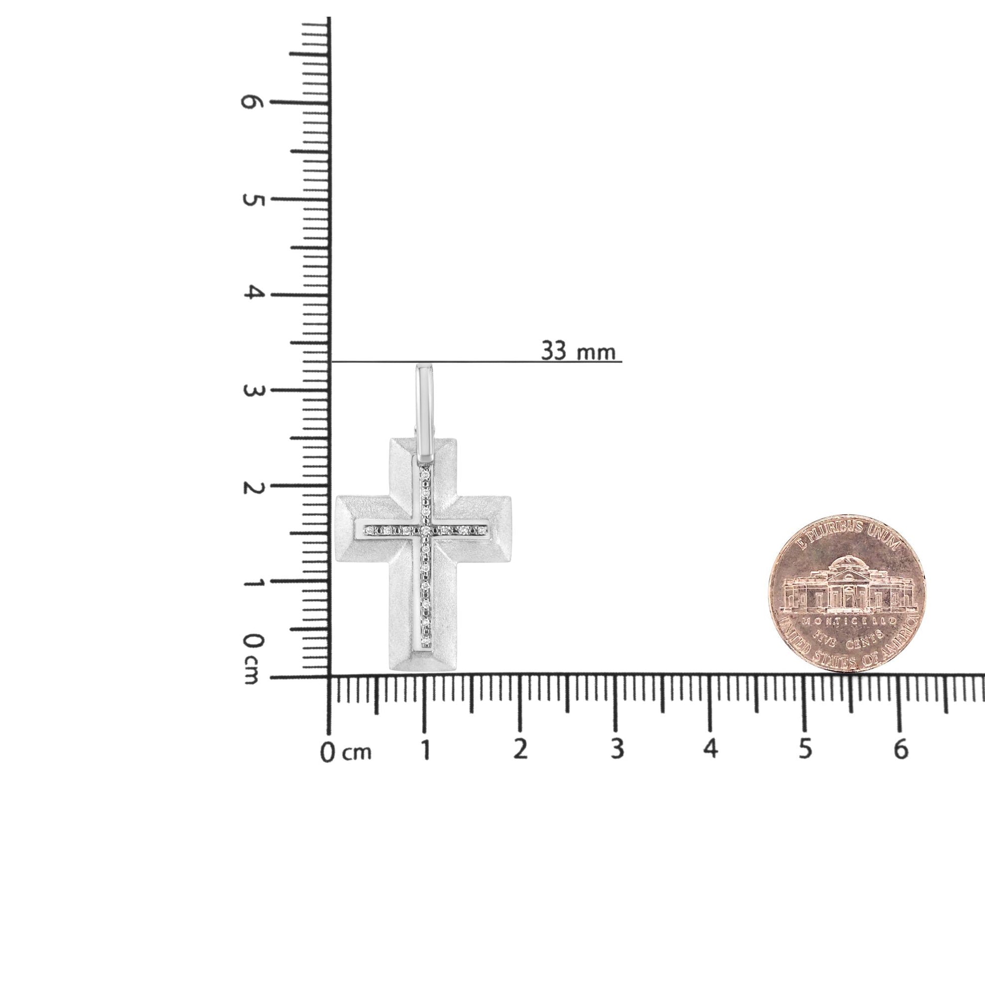 White cross-shaped necklace pendant with a vertical and horizontal row of white round gemstone in prong setting, placed flat on a ruler and scale with a coin for size reference, shot from top angle.