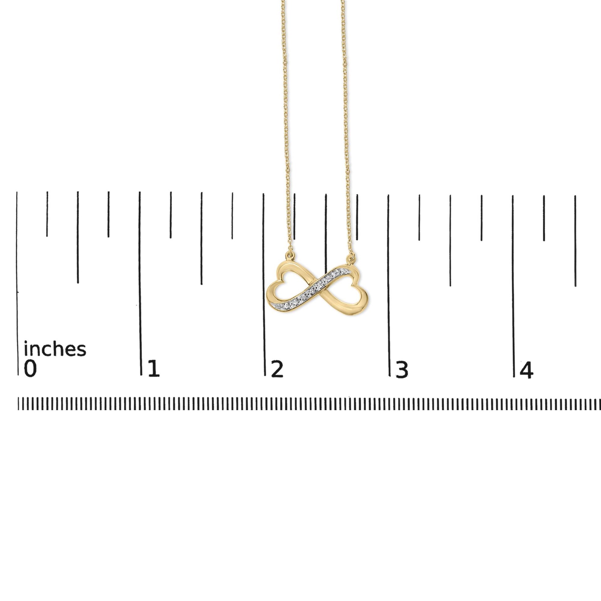 Close-up front view of a yellow metal infinity necklace pendant with two heart shapes and a diagonal row of white gemstones in pave setting, shown on a measurement scale.