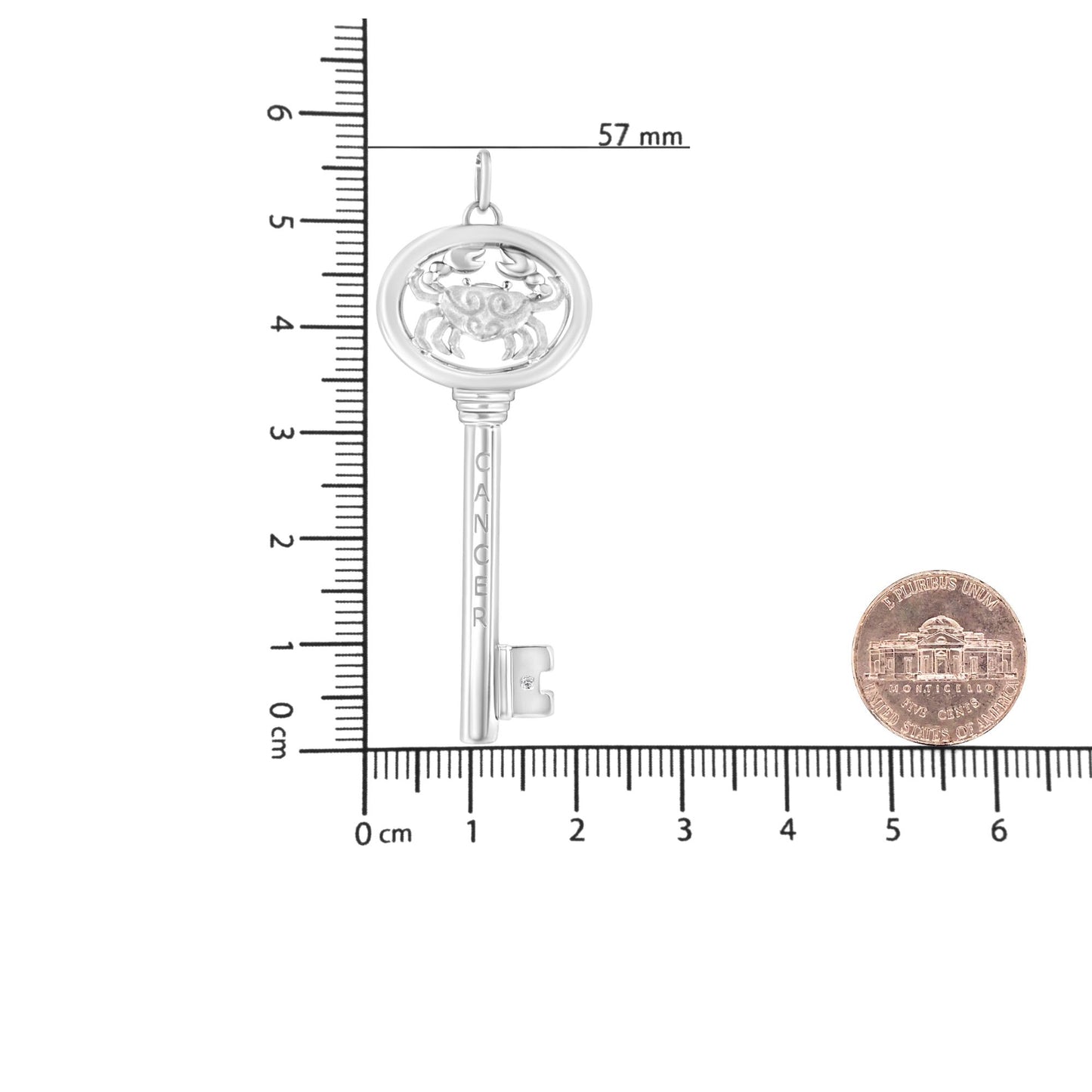 White metal necklace pendant in the shape of a key with a crab design at the top and an engraved word on the shaft, featuring a single round brownish gemstone in bezel setting near the bottom, photographed flat on a ruler and scale for size reference from a top-down angle.