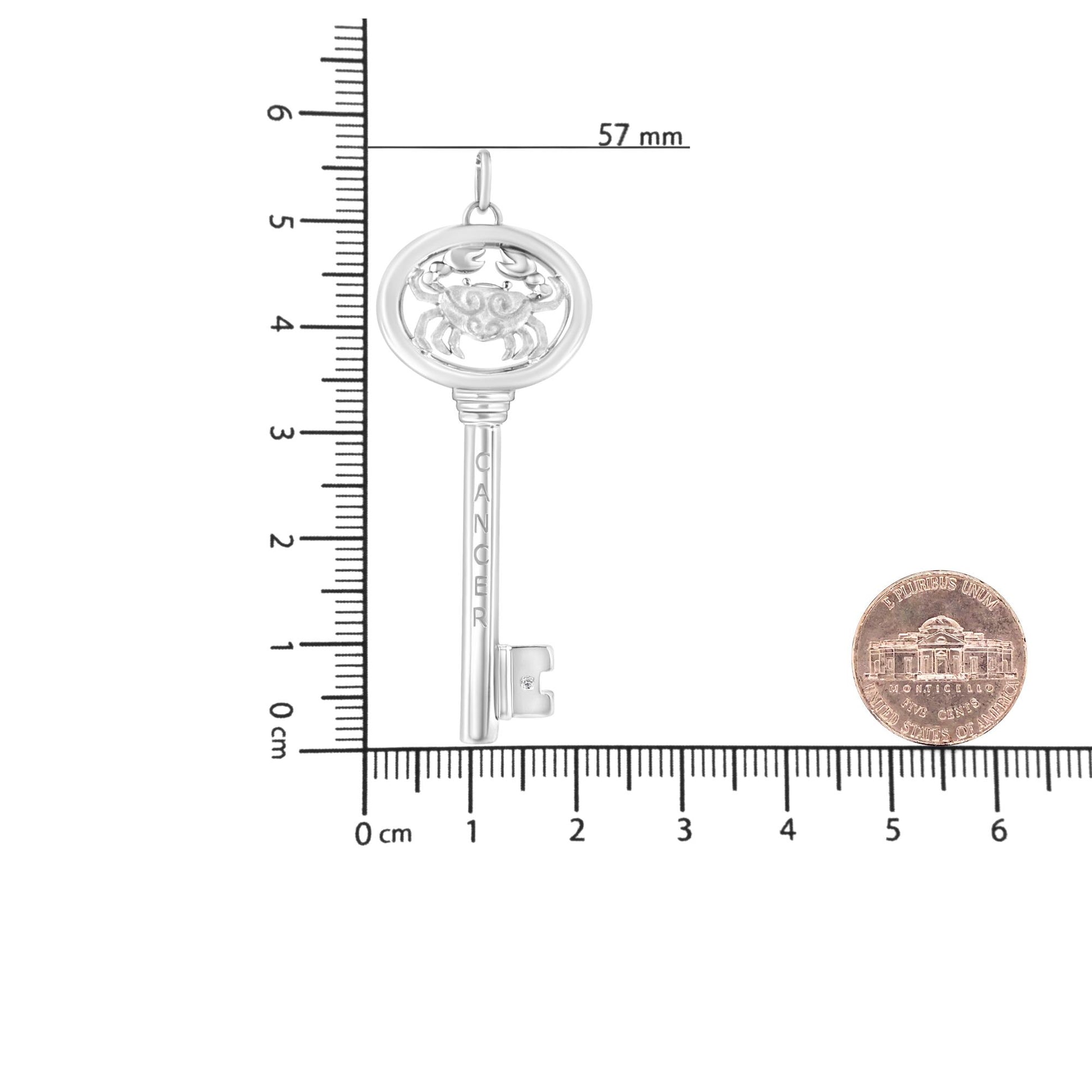 White metal necklace pendant in the shape of a key with a crab design at the top and an engraved word on the shaft, featuring a single round brownish gemstone in bezel setting near the bottom, photographed flat on a ruler and scale for size reference from a top-down angle.