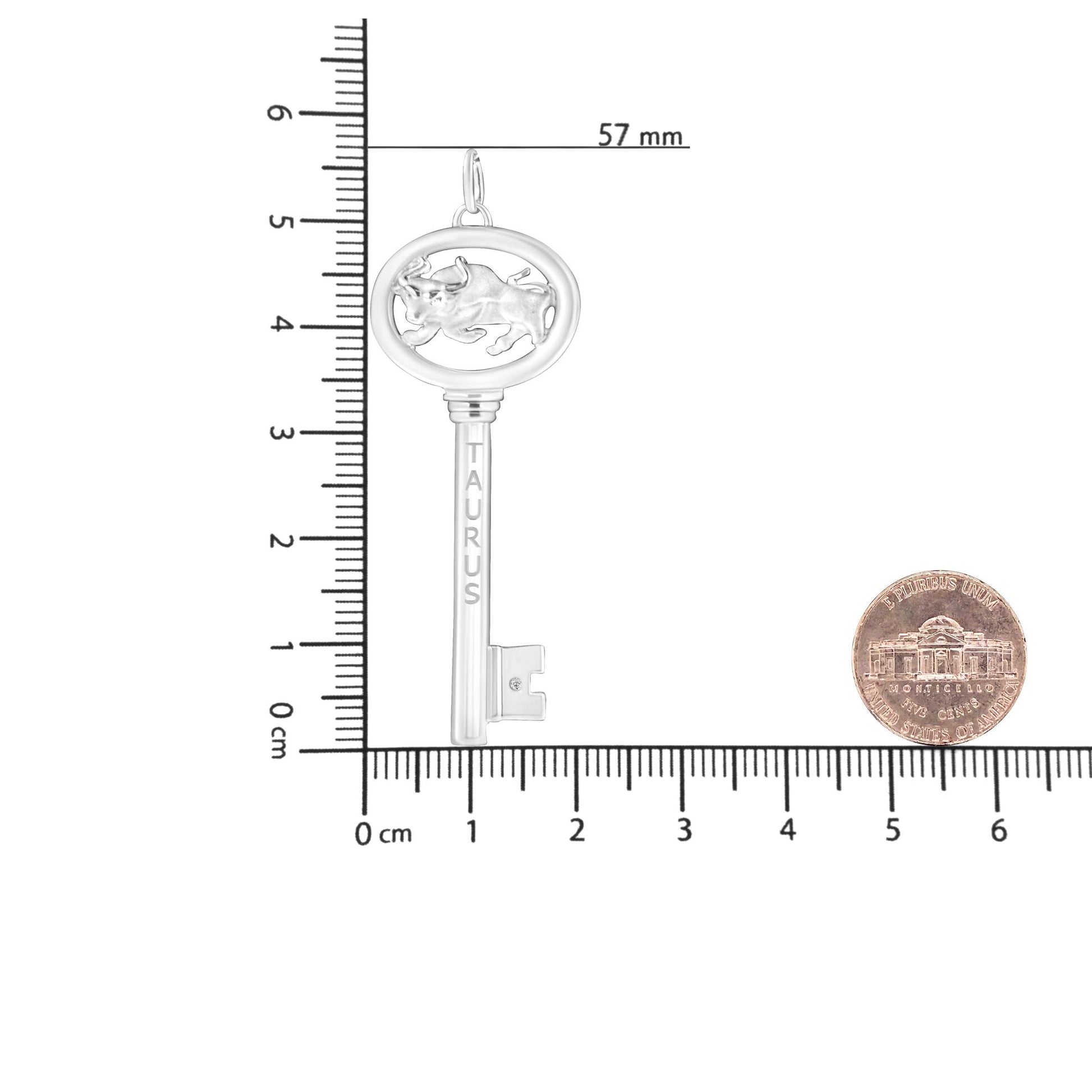 White metal key-shaped necklace pendant with a raised bull design and engraved word 'TAURUS', featuring a round brownish gemstone in bezel setting; shown flat on a ruler and coin for size reference, top-down angle.