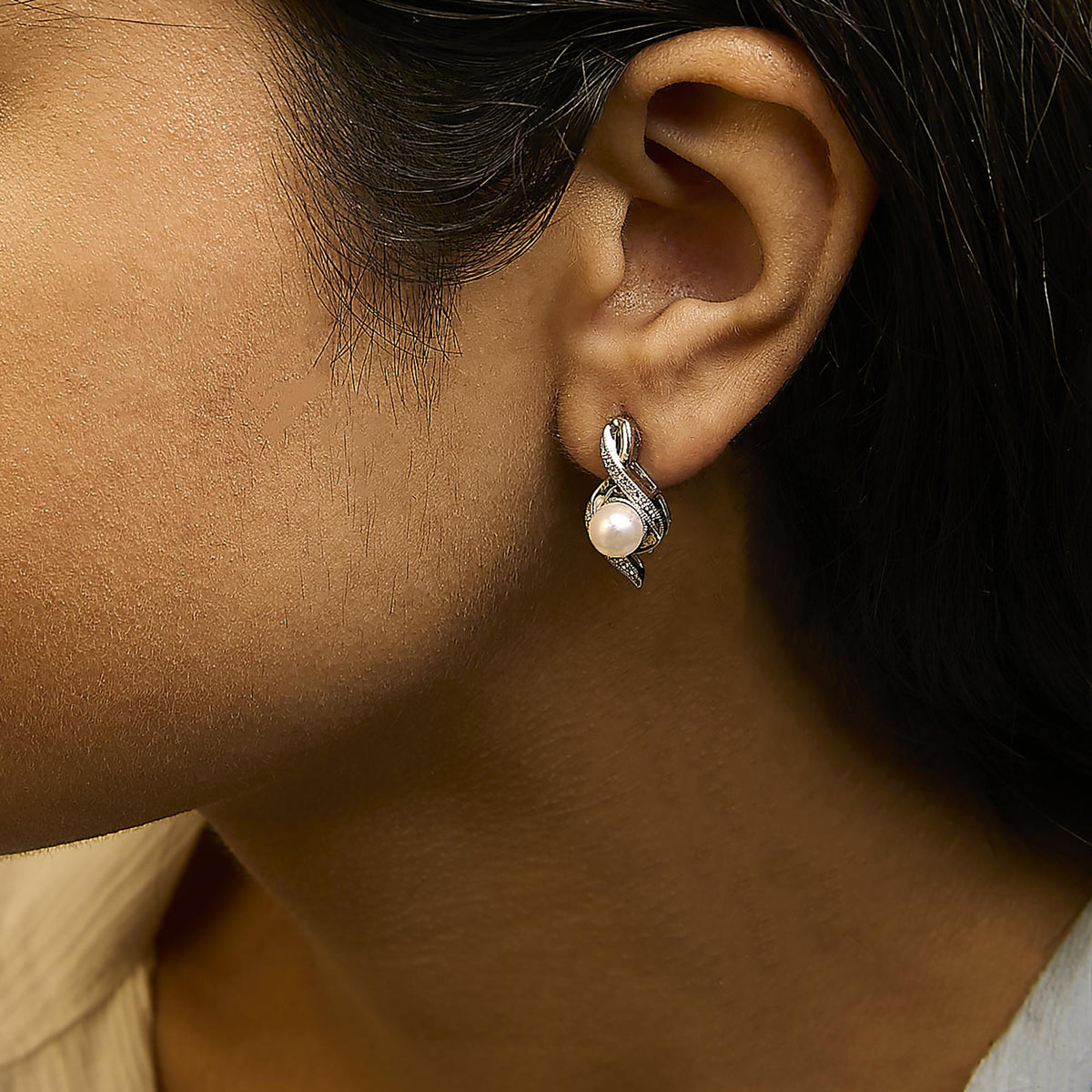 Close-up side view of a model wearing white metal earrings with a twisting design, adorned with white round gemstones in a pave setting and a single large white pearl at the bottom.