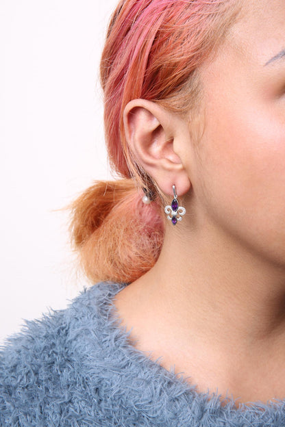 Close-up side view of a model wearing white metal earrings featuring two purple gemstones and multiple small white round gemstones in prong settings.