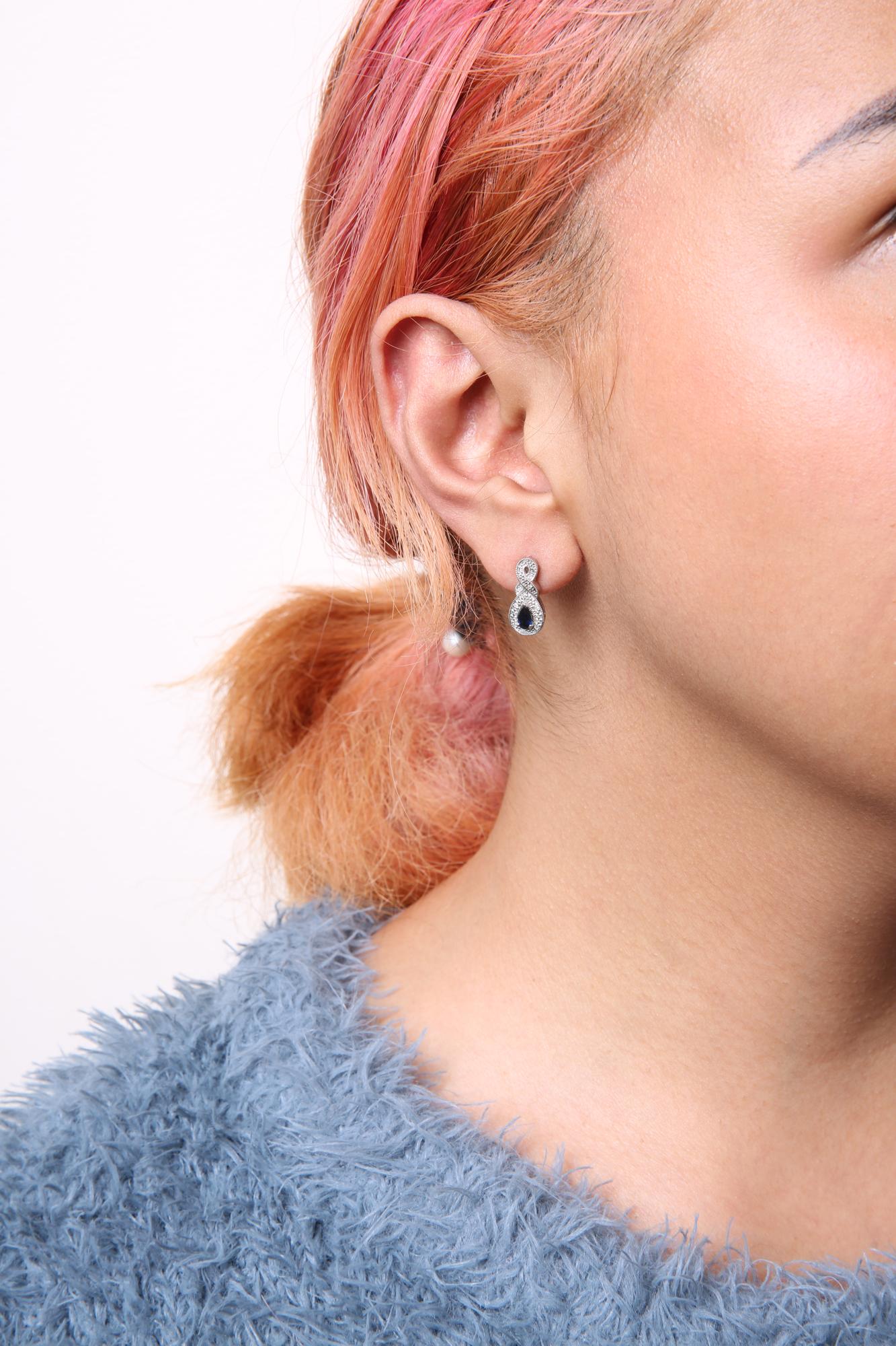 Close-up side view of white metal earrings on a model's ear featuring an infinity shape design with small round white gemstones and a pear-shaped blue gemstone at the bottom.