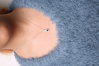 Front view of a white metal necklace with a pendant featuring a pear-shaped blue gemstone surrounded by round white gemstones, worn on a model with a blue fuzzy top.