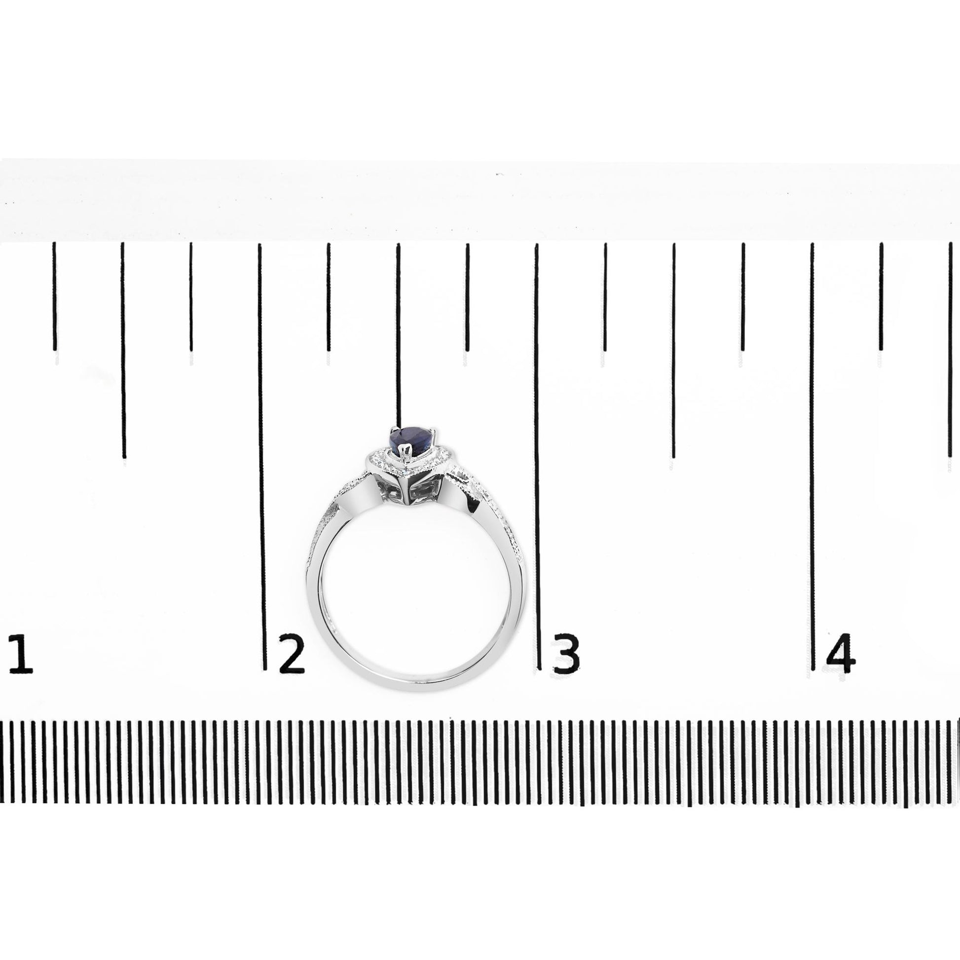 White metal ring with a pear-shaped blue gemstone and round white gemstones in prong settings, shown from a top angle on a ruler for size reference.