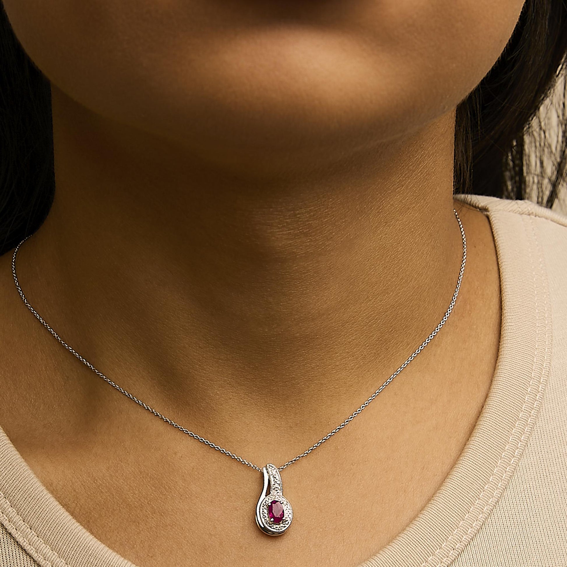 Close-up image showing a white metal necklace with a teardrop-shaped pendant worn on a model, featuring a red oval gemstone surrounded by small white round gemstones in a pave setting.
