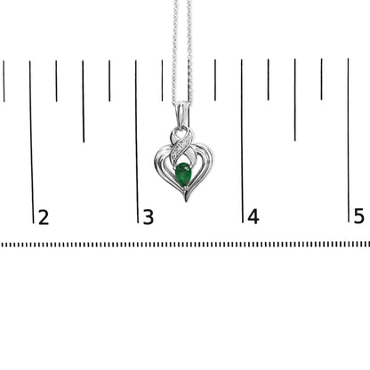 Close-up front view of a white metal heart-shaped necklace pendant with a pear-shaped green gemstone in the center and four round white gemstones above it, shown hanging on a chain against a ruler for size reference.