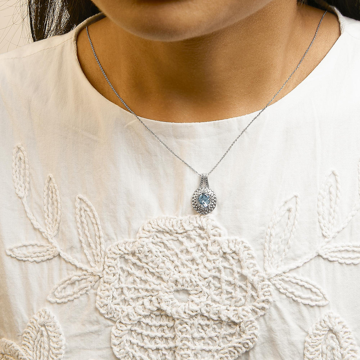 White metal necklace with a round pave setting of white gemstones surrounding a blue oval gemstone pendant, shown worn on a model from a front angle.