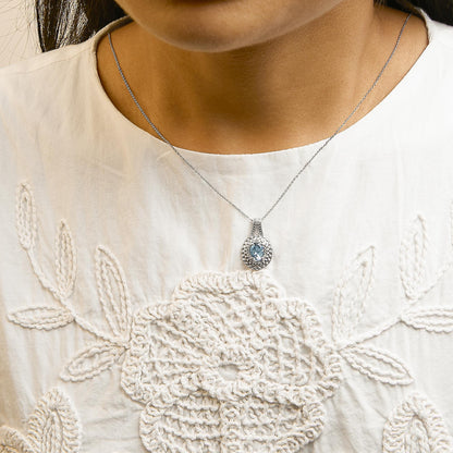 White metal necklace with a round pave setting of white gemstones surrounding a blue oval gemstone pendant, shown worn on a model from a front angle.