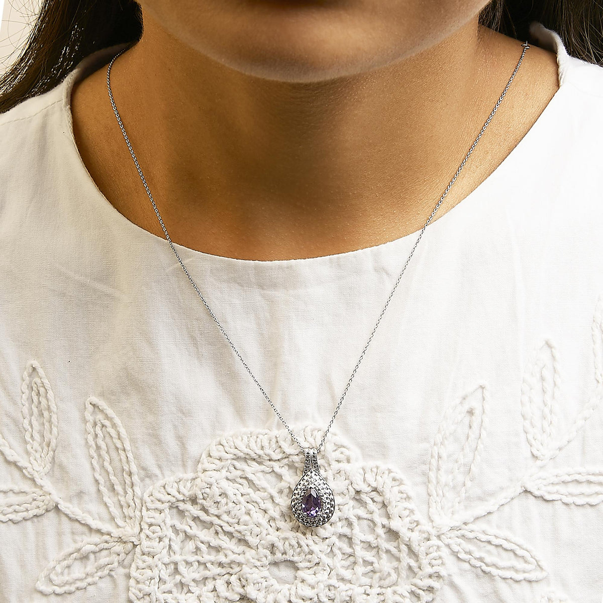 White metal necklace with a pear-shaped purple gemstone pendant surrounded by pave-set white round gemstones, shown worn on a model in a front close-up angle.