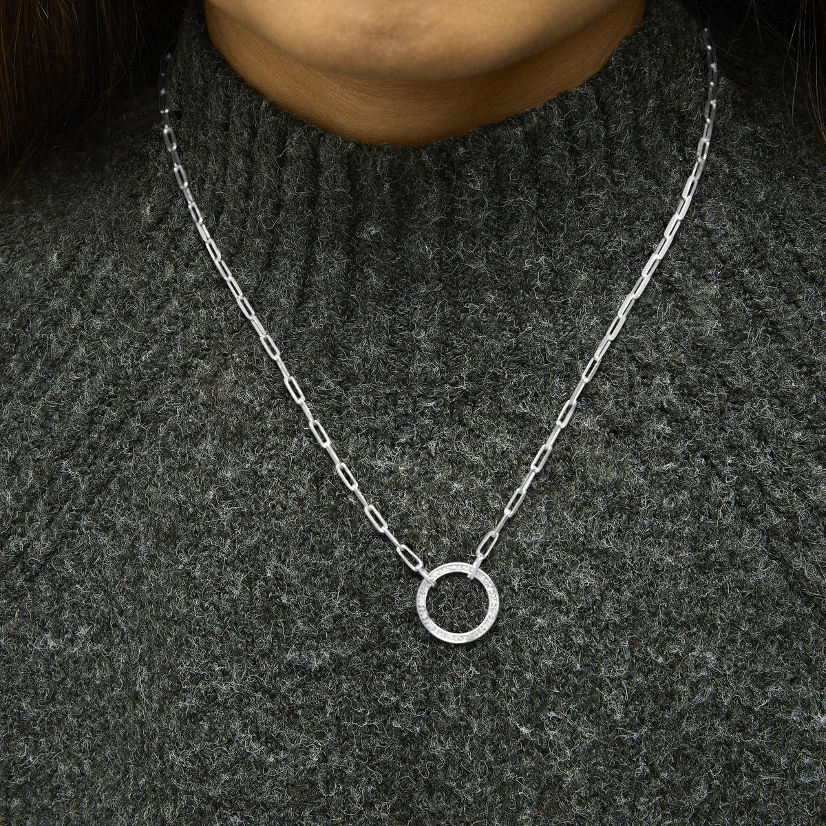 Close-up front view of a model wearing a white metal necklace with a circular pendant featuring pave-set white gemstones, shown against a dark textured top.